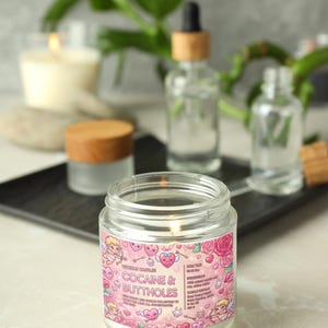 May include: A lit jar candle with a pink label that reads "Cocaine & Buttholes" with cartoon graphics. The candle sits on a black tray with other glass bottles and a lit candle in the background. The candle is white.