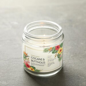 May include: A lit, clear glass jar candle with a label that reads "COCAINE & BUTTHOLES." The label is decorated with tropical flowers and foliage. The candle is on a grey surface.