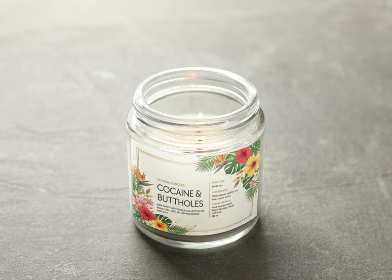 May include: A lit, clear glass jar candle with a label that reads "COCAINE & BUTTHOLES." The label is decorated with tropical flowers and foliage. The candle is on a grey surface.