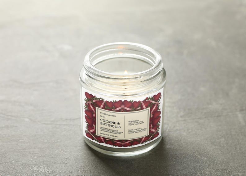 May include: A clear glass jar candle with a lit wick. The label features a strawberry and ribbon design with the text "COCAINE & BUTTHOLES". The candle is on a gray surface.