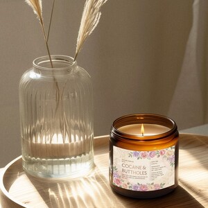 May include: A lit candle in a brown glass jar with a floral label that reads "Cocaine & Buttholes" sits on a wooden tray. A clear glass vase with dried plants is to the left. The candle's flame glows brightly.