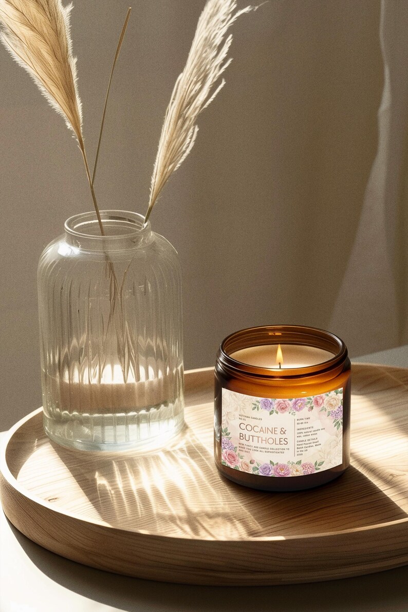 May include: A lit candle in a brown glass jar with a floral label that reads "Cocaine & Buttholes" sits on a wooden tray. A clear glass vase with dried plants is to the left. The candle's flame glows brightly.