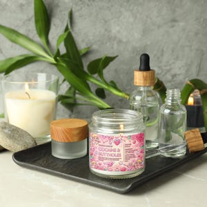 May include: A lit candle in a glass jar with a wooden lid, labeled "COCAINE & BUTTHOLES" with a pink and white label. The candle sits on a black tray with other glass bottles and a lit candle in a glass. Green bamboo stalks are in the background.