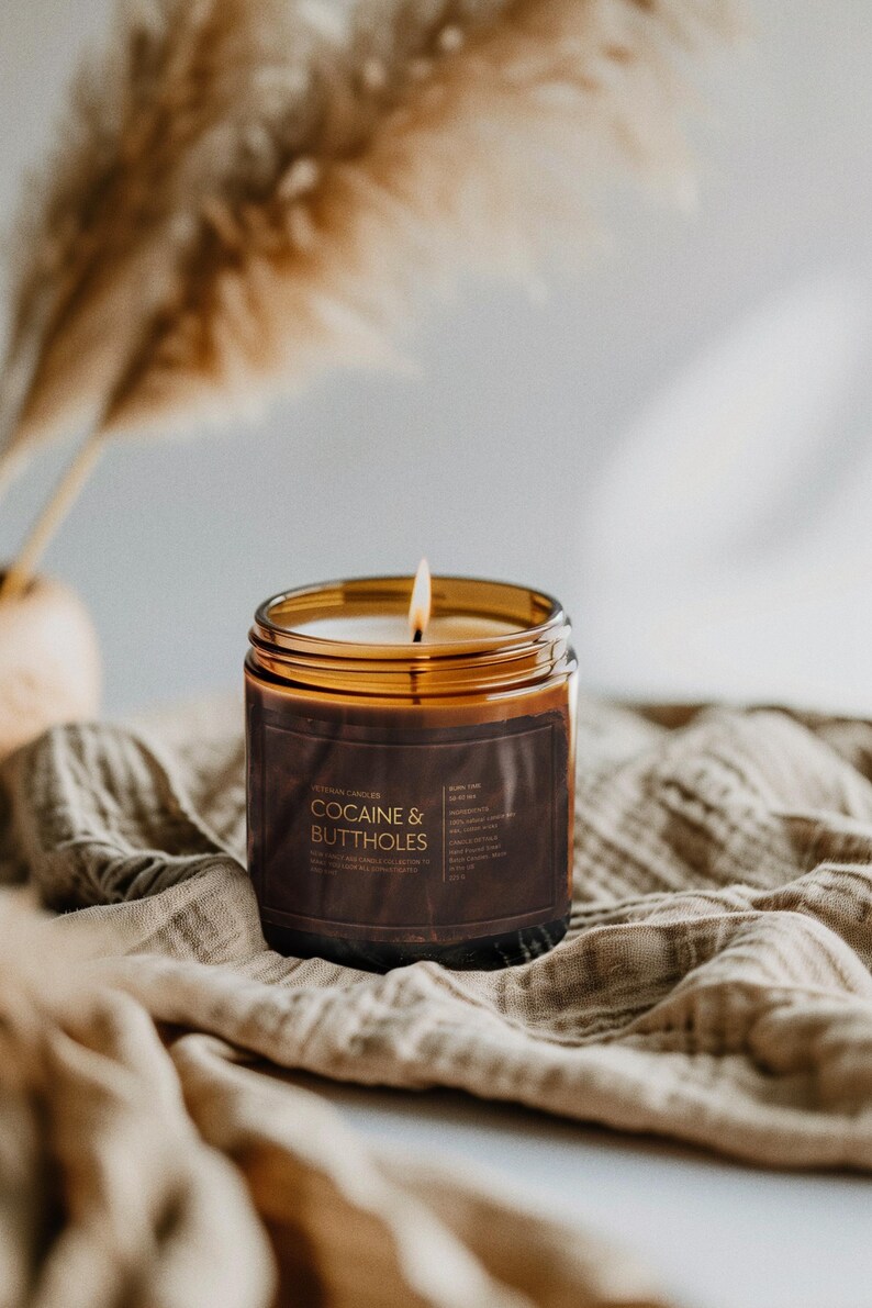 May include: A lit candle in a brown glass jar with a dark brown label that reads "COCAINE & BUTTHOLES". The candle sits on a beige cloth with dried ornamental grass in the background. The candle's flame is lit.