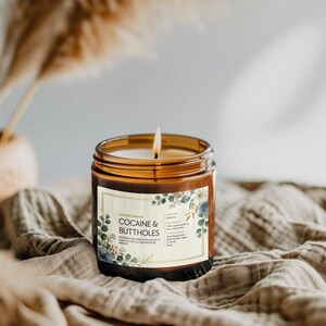 May include: A lit candle in a brown glass jar with a label that reads "COCAINE & BUTTHOLES". The label has a floral design with blue and green accents. The candle sits on a tan cloth.