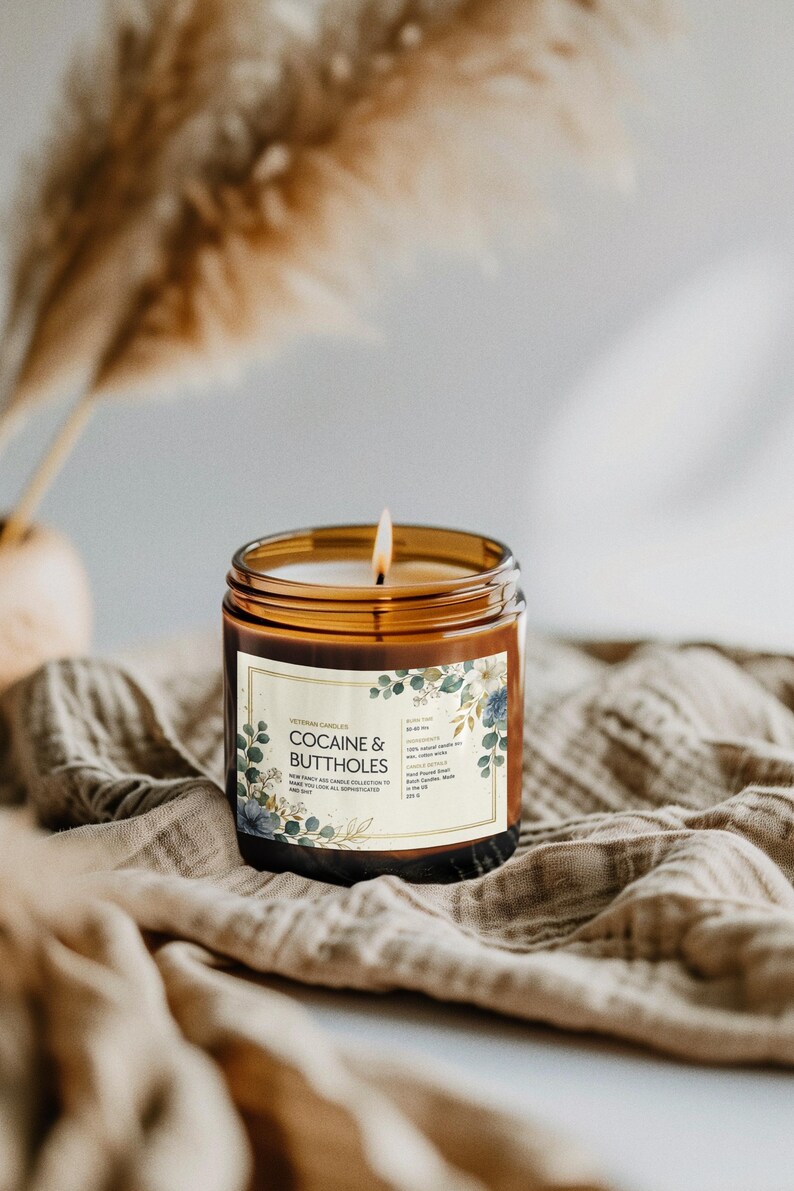 May include: A lit candle in a brown glass jar with a label that reads "COCAINE & BUTTHOLES". The label has a floral design with blue and green accents. The candle sits on a tan cloth.