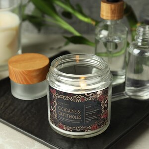 May include: A lit candle in a clear glass jar with a wooden lid, labeled "COCAINE & BUTTHOLES" in a decorative frame. Other glass bottles and a lit candle are in the background, all on a black tray.