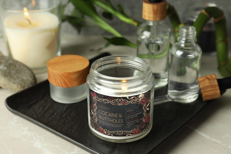 May include: A lit candle in a clear glass jar with a wooden lid, labeled "COCAINE & BUTTHOLES" in a decorative frame. Other glass bottles and a lit candle are in the background, all on a black tray.