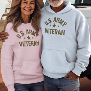 Puede incluir: Una pareja con sudaderas a juego con el texto "U.S. ARMY VETERAN" en verde oliva. Una sudadera es rosa claro, la otra azul claro. Ambas tienen un bolsillo delantero y se combinan con vaqueros y sombreros de vaquero. Tema militar.