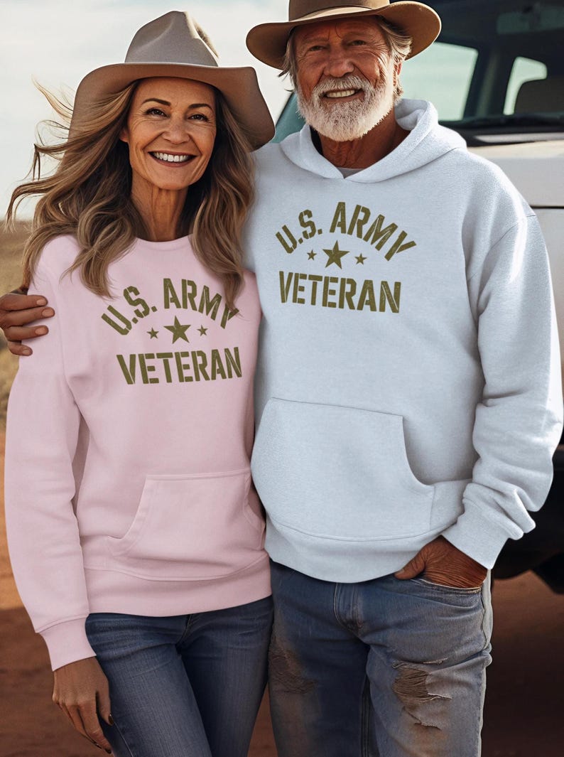 Puede incluir: Una pareja con sudaderas a juego con el texto "U.S. ARMY VETERAN" en verde oliva. Una sudadera es rosa claro, la otra azul claro. Ambas tienen un bolsillo delantero y se combinan con vaqueros y sombreros de vaquero. Tema militar.