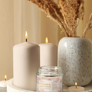 Puede incluir: Una naturaleza muerta con velas encendidas y un jarr&oacute;n decorativo. La pieza central es una vela de tarro de vidrio con el texto "COCAINE & BUTTHOLES" rodeado de un dise&ntilde;o floral. Las velas son blancas y est&aacute;n sobre una bandeja moteada.