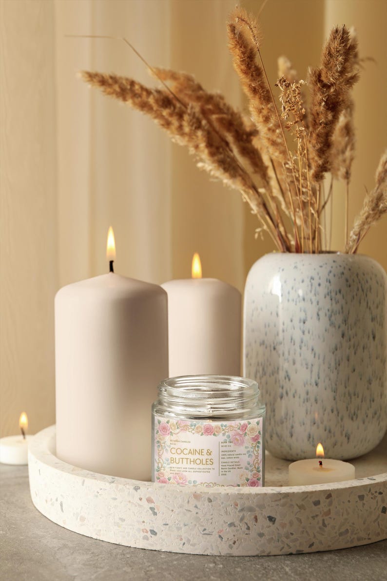Puede incluir: Una naturaleza muerta con velas encendidas y un jarr&oacute;n decorativo. La pieza central es una vela de tarro de vidrio con el texto "COCAINE & BUTTHOLES" rodeado de un dise&ntilde;o floral. Las velas son blancas y est&aacute;n sobre una bandeja moteada.