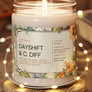May include: A lit, pale pink candle in a glass jar with a floral label. The label reads "DAYSHIFT & C. DIFF" and "NEW FANCY ASS NURSE CANDLE COLLECTION". The candle sits on a wooden coaster, surrounded by string lights. Burn time is 45-50 hours.