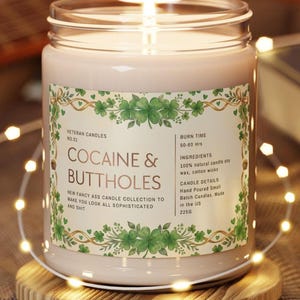 Op de afbeelding: Een brandende kaars in een glazen pot met een etiket met de tekst "COCAINE & BUTTHOLES". Het etiket heeft een groen klaverontwerp en de tekst "Veteran Candles". De kaars staat op een houten onderzetter en is omgeven door lichtslingers. De kaars weegt 225g.