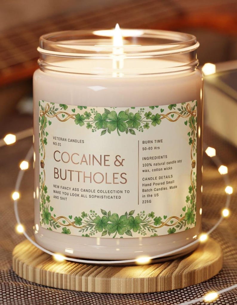 Op de afbeelding: Een brandende kaars in een glazen pot met een etiket met de tekst "COCAINE & BUTTHOLES". Het etiket heeft een groen klaverontwerp en de tekst "Veteran Candles". De kaars staat op een houten onderzetter en is omgeven door lichtslingers. De kaars weegt 225g.