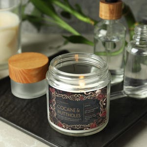 Pu&ograve; includere: Una candela accesa in un barattolo di vetro trasparente con coperchio in legno, etichettata "COCAINE & BUTTHOLES" in una cornice decorativa. Altre bottiglie di vetro e una candela accesa sono sullo sfondo, il tutto su un vassoio nero.
