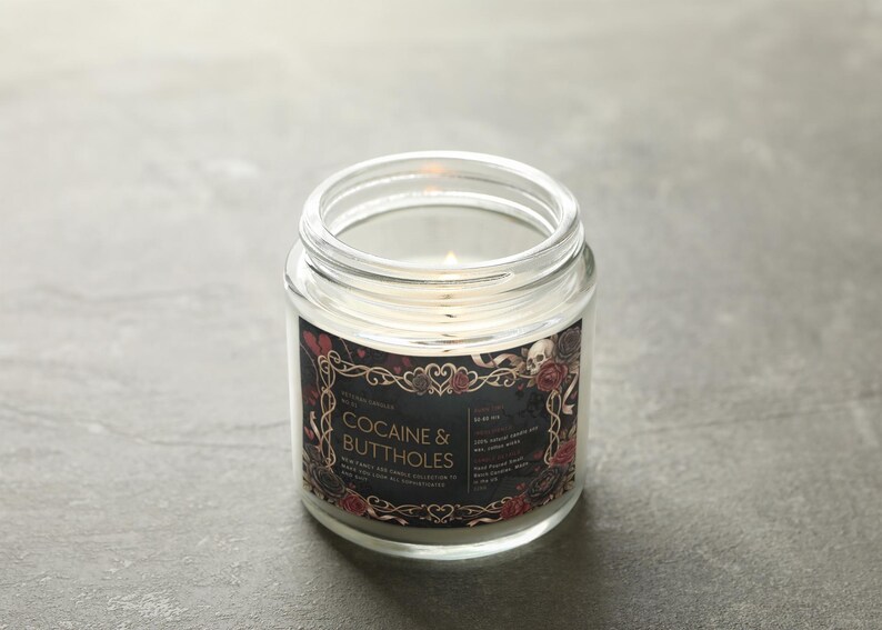 May include: A lit, clear glass jar candle with a black label featuring the text "COCAINE & BUTTHOLES" in white. The label is decorated with red roses, skulls, and ornate gold-colored scrollwork. The candle sits on a gray surface.