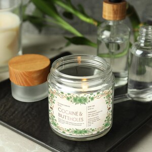 Op de afbeelding: Een brandende glazen potkaars met de tekst "Cocaine & Buttholes" op een label met groene accenten. Andere items zijn een kleine pot met een houten deksel, twee heldere flessen en een brandende kaars in een glas.