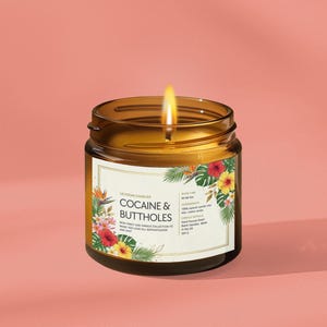 Może przedstawiać: Zapalonego świeca w bursztynowym szklanym słoiku z etykietą z napisem "COCAINE & BUTTHOLES". Etykieta jest ozdobiona tropikalnymi kwiatami i liśćmi. Świeca stoi na r&oacute;żowym tle.