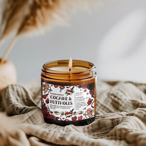 Puede incluir: Una vela encendida en un frasco de vidrio marr&oacute;n con una etiqueta que dice "COCAINE & BUTTHOLES". La etiqueta presenta ilustraciones de estilo tatuaje de corazones, rosas y querubines. La vela est&aacute; sobre una tela beige.