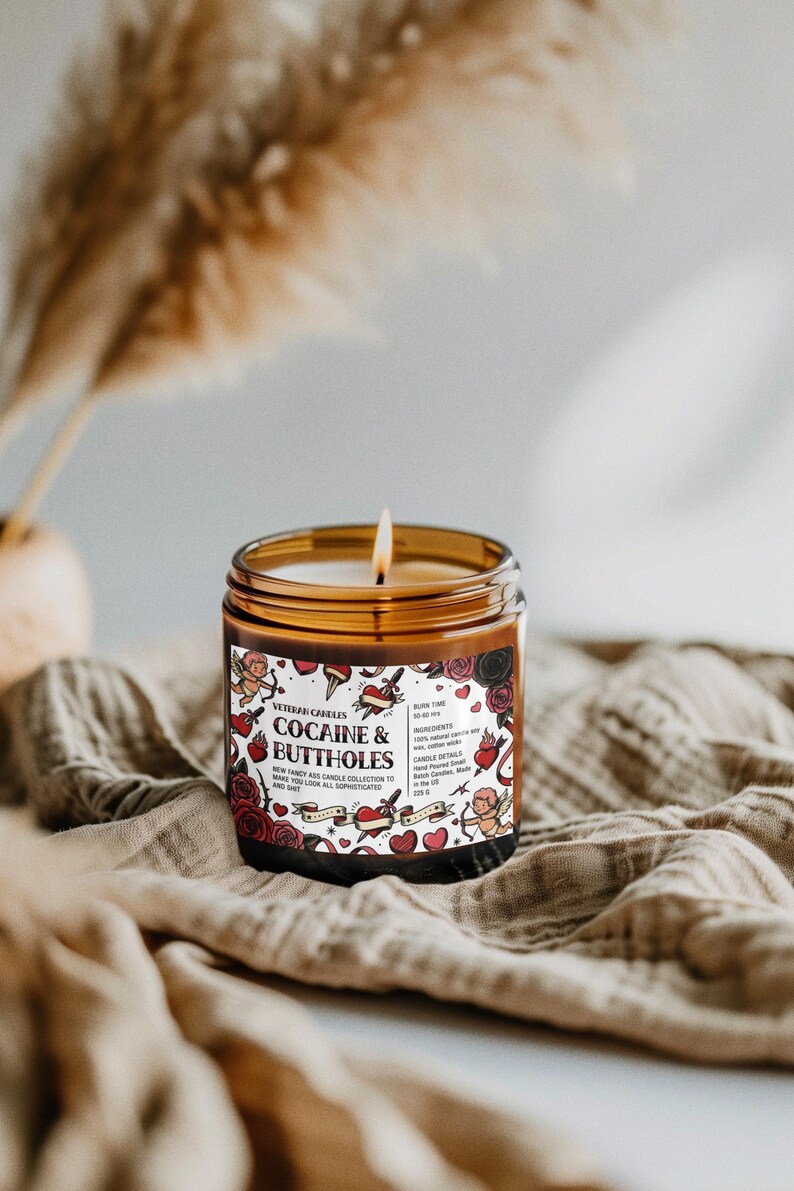 Puede incluir: Una vela encendida en un frasco de vidrio marr&oacute;n con una etiqueta que dice "COCAINE & BUTTHOLES". La etiqueta presenta ilustraciones de estilo tatuaje de corazones, rosas y querubines. La vela est&aacute; sobre una tela beige.