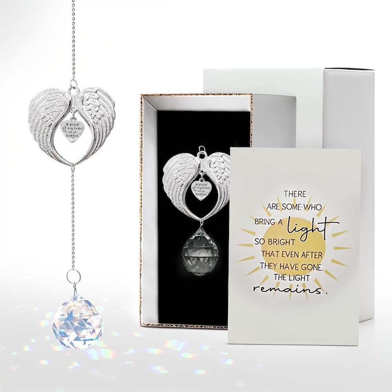 May include: A silver angel wing ornament with a heart-shaped charm that reads "A piece of my heart is in heaven." A faceted crystal hangs below. The ornament is displayed in a gift box with a card that reads "There are some who bring a light so bright that even after they have gone, the light remains."