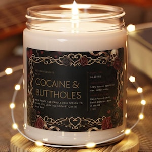 May include: A lit, white candle in a glass jar with a black label featuring the text "COCAINE & BUTTHOLES". The label includes floral and skull motifs. The candle sits on a wooden coaster, surrounded by string lights.