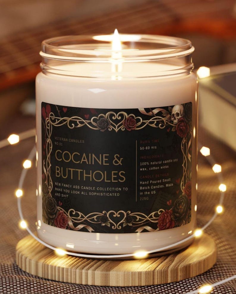 May include: A lit, white candle in a glass jar with a black label featuring the text "COCAINE & BUTTHOLES". The label includes floral and skull motifs. The candle sits on a wooden coaster, surrounded by string lights.