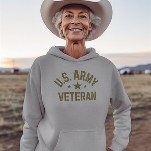 Puede incluir: Una sudadera gris claro con el texto "U.S. ARMY VETERAN" en verde oliva. La persona de la imagen lleva un sombrero de vaquero blanco. El fondo es una escena borrosa al aire libre.