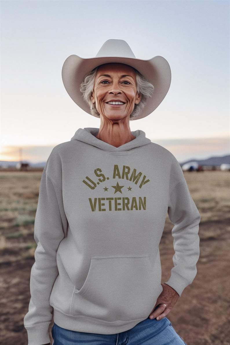 Puede incluir: Una sudadera gris claro con el texto "U.S. ARMY VETERAN" en verde oliva. La persona de la imagen lleva un sombrero de vaquero blanco. El fondo es una escena borrosa al aire libre.