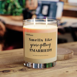 May include: A clear glass jar candle with a light orange and yellow ombre wax. The label reads "Smells like you're getting *MARRIED!* Soy Candle. Burning time: 50-60 hours."