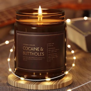 May include: A lit brown glass jar candle with the text "COCAINE & BUTTHOLES" on a brown label. The candle sits on a wooden coaster, surrounded by string lights. The candle's label also includes the text "Veteran Candles".