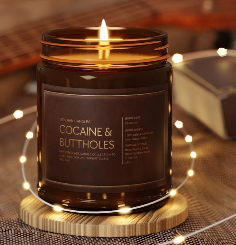May include: A lit brown glass jar candle with the text "COCAINE & BUTTHOLES" on a brown label. The candle sits on a wooden coaster, surrounded by string lights. The candle's label also includes the text "Veteran Candles".