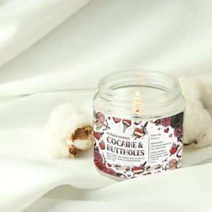 May include: A clear glass jar candle with a lit wick, labeled "Cocaine & Buttholes." The label features tattoo-style illustrations of roses, hearts, and cherubs. The candle is set on a white fabric with cotton buds.