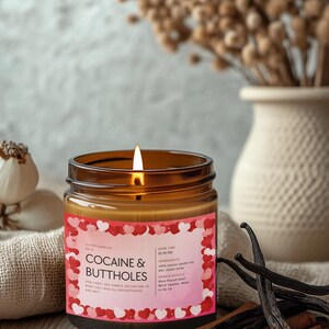May include: A lit candle in a brown glass jar with a pink label that reads "COCAINE & BUTTHOLES." The label is decorated with red and pink hearts. Vanilla beans and cinnamon sticks are arranged on a wooden surface.