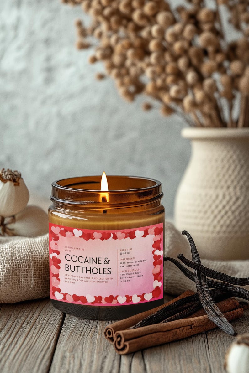 May include: A lit candle in a brown glass jar with a pink label that reads "COCAINE & BUTTHOLES." The label is decorated with red and pink hearts. Vanilla beans and cinnamon sticks are arranged on a wooden surface.