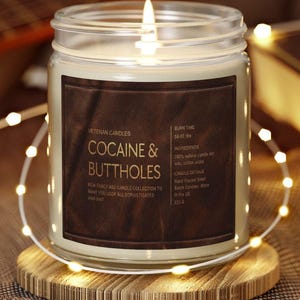 May include: A lit candle in a glass jar with a brown label that reads "COCAINE & BUTTHOLES." The candle sits on a wooden coaster and is surrounded by string lights. The label also states "New fancy ass candle collection to make you look all sophisticated and shit."