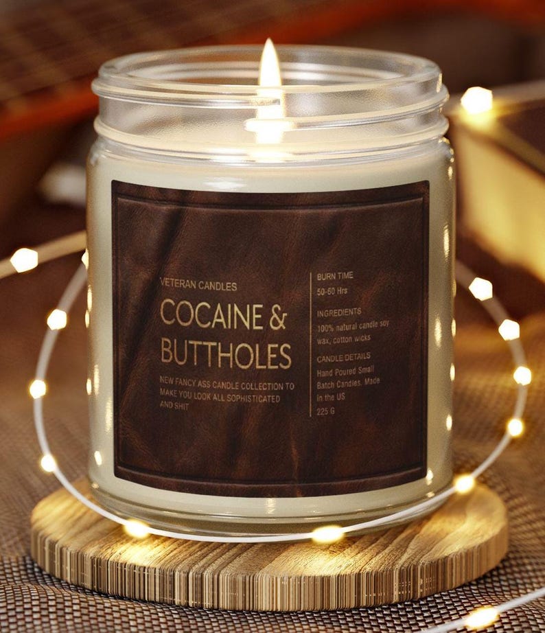 May include: A lit candle in a glass jar with a brown label that reads "COCAINE & BUTTHOLES." The candle sits on a wooden coaster and is surrounded by string lights. The label also states "New fancy ass candle collection to make you look all sophisticated and shit."