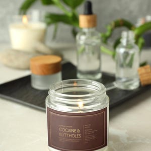 May include: A lit glass jar candle with a brown label that reads "Cocaine & Buttholes" in white text. Other candles and bottles are in the background. The candle is on a white surface.