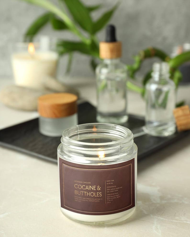 May include: A lit glass jar candle with a brown label that reads "Cocaine & Buttholes" in white text. Other candles and bottles are in the background. The candle is on a white surface.