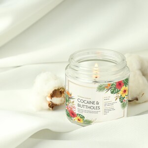 May include: A lit glass jar candle with the text "COCAINE & BUTTHOLES" on the label, decorated with flowers. The candle sits on white fabric, with cotton bolls nearby.