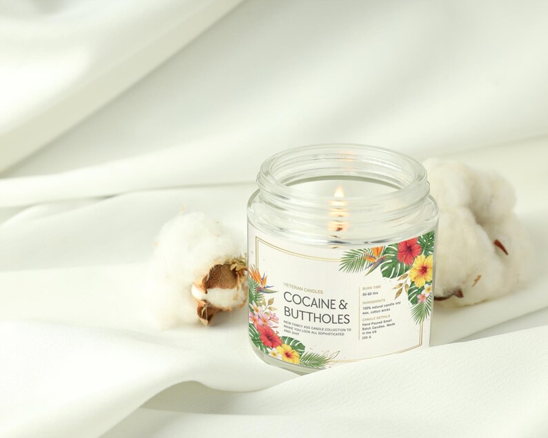 May include: A lit glass jar candle with the text "COCAINE & BUTTHOLES" on the label, decorated with flowers. The candle sits on white fabric, with cotton bolls nearby.
