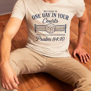 May include: A beige t-shirt with the text "Better is One Day in Your Courts than a Thousand Elsewhere Psalm 84:10" in navy blue. The shirt features a volleyball net graphic. The person is wearing khaki pants and sitting on a wooden floor.