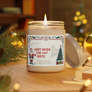 May include: A white candle jar with a red and green Christmas-themed label that reads "Light When The Cat Shits". The label also includes a cartoon Santa Claus and a Christmas tree.
