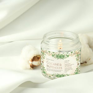Może przedstawiać: Zapalone, przezroczyste świeca w szklanym słoiku z etykietą z napisem "COCAINE & BUTTHOLES" otoczoną zielonymi kwiatowymi wzorami. Świeca stoi na białej tkaninie obok kulek bawełny. Świeca jest częścią nowej, fantazyjnej kolekcji świec.
