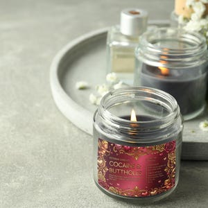 Peut inclure: Une bougie grise fonc&eacute;e allum&eacute;e dans un bocal en verre transparent avec une &eacute;tiquette qui dit "Cocaine & Buttholes". L'&eacute;tiquette a un design rouge et or avec des roses et des ch&eacute;rubins. D'autres bougies et un plateau sont en arri&egrave;re-plan.