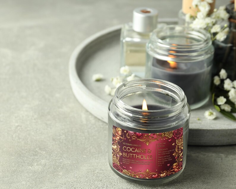 Peut inclure: Une bougie grise fonc&eacute;e allum&eacute;e dans un bocal en verre transparent avec une &eacute;tiquette qui dit "Cocaine & Buttholes". L'&eacute;tiquette a un design rouge et or avec des roses et des ch&eacute;rubins. D'autres bougies et un plateau sont en arri&egrave;re-plan.