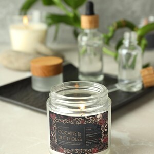 Pu&ograve; includere: Una candela accesa in un barattolo di vetro trasparente con un'etichetta che recita "Cocaine & Buttholes". L'etichetta ha uno sfondo nero con rose rosse e un disegno di teschio. Altre bottiglie di vetro e una candela sono sullo sfondo.