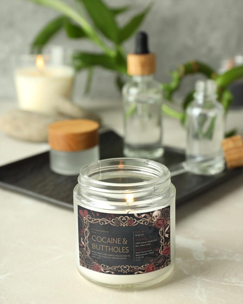 Pu&ograve; includere: Una candela accesa in un barattolo di vetro trasparente con un'etichetta che recita "Cocaine & Buttholes". L'etichetta ha uno sfondo nero con rose rosse e un disegno di teschio. Altre bottiglie di vetro e una candela sono sullo sfondo.