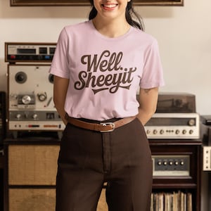 May include: A light pink t-shirt with the phrase "Well... Sheeyit" in brown lettering. The person is wearing brown pants and a brown belt. The background includes vintage audio equipment and a shelf with decorative items.